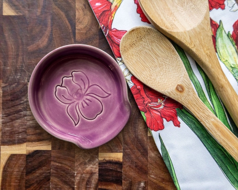 A purple ceramic spoon rest with a flower design sits on a wooden countertop next to two wooden spoons and a floral-patterned cloth.