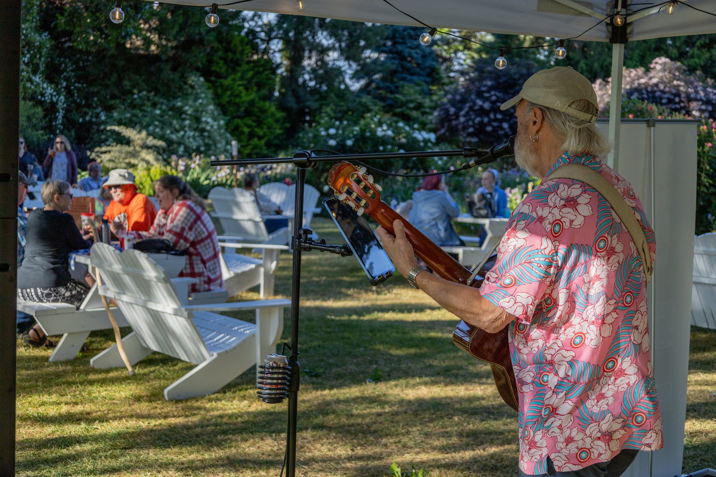 A man with gray hair, wearing a pink floral shirt and a tan baseball cap, plays a guitar under a canopy strung with lights. Audience members sit in white chairs in the background.
