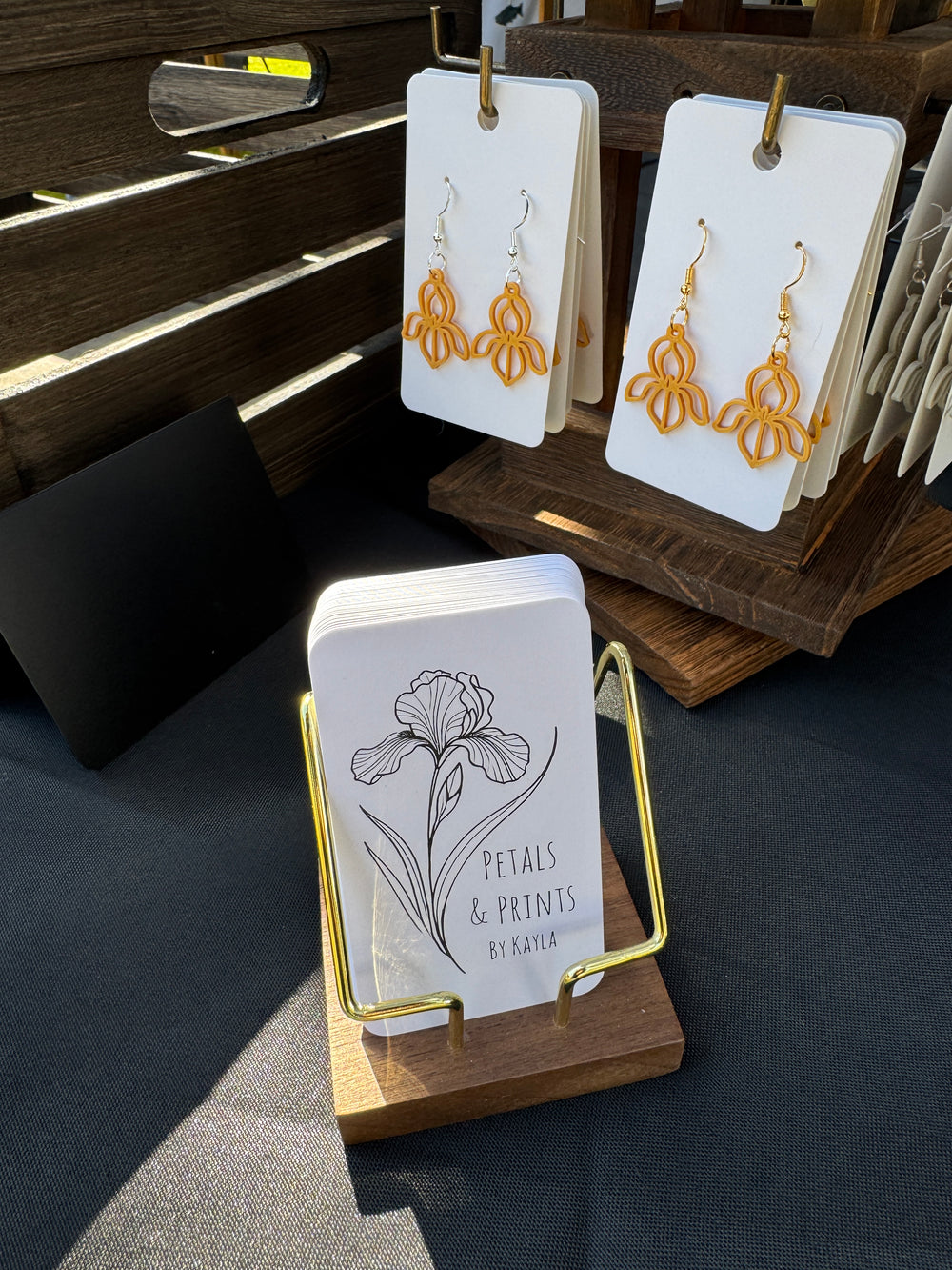 Display of yellow earrings on white cards and a stack of business cards labeled Petals & Prints by Kayla with a flower illustration, set on a wooden stand at an outdoor market.
