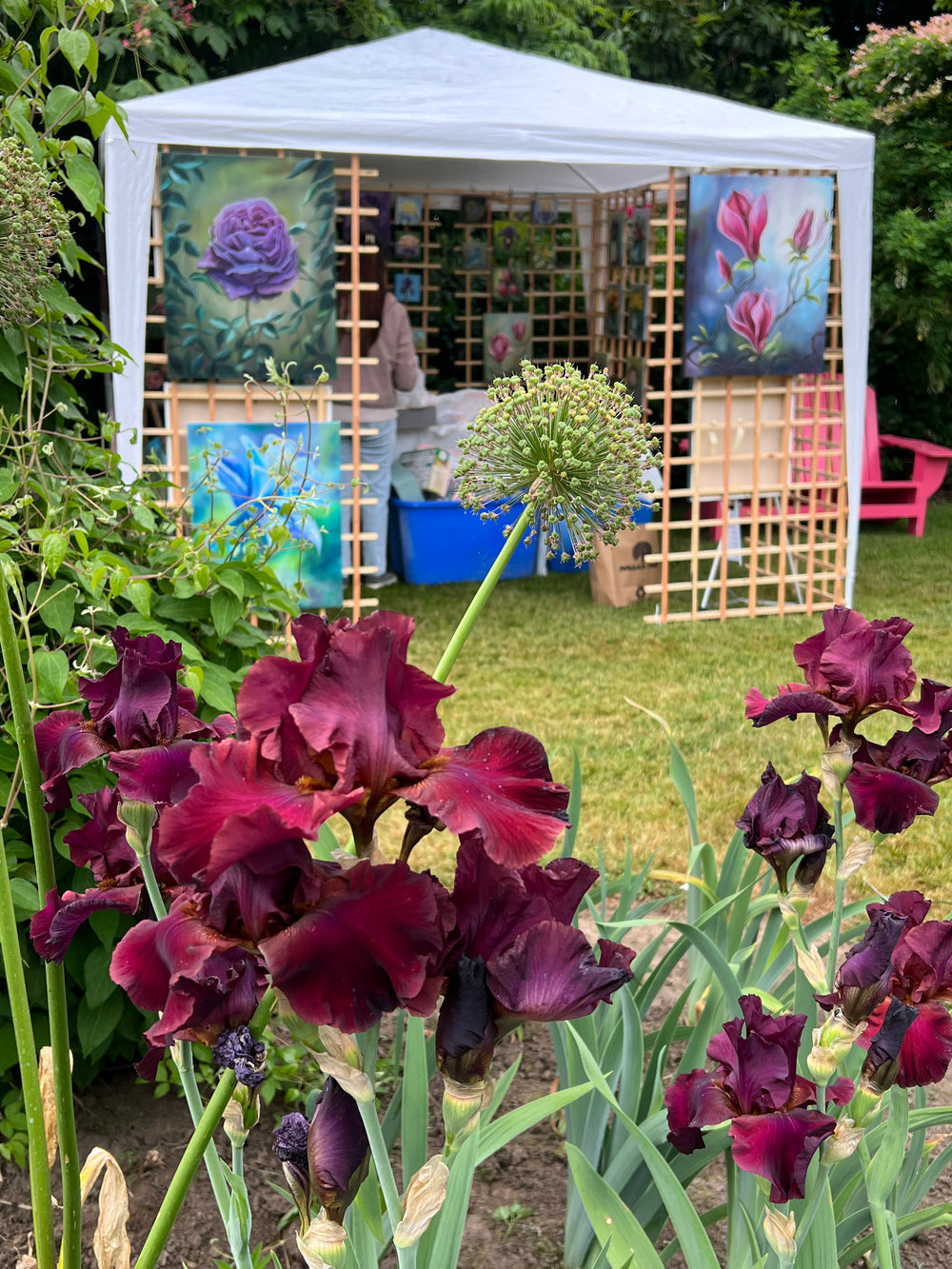 Outdoor art booth with colorful flower paintings displayed on a wooden grid. In the foreground, dark purple irises and other plants bloom in a garden, while a person inside the booth is partially visible.